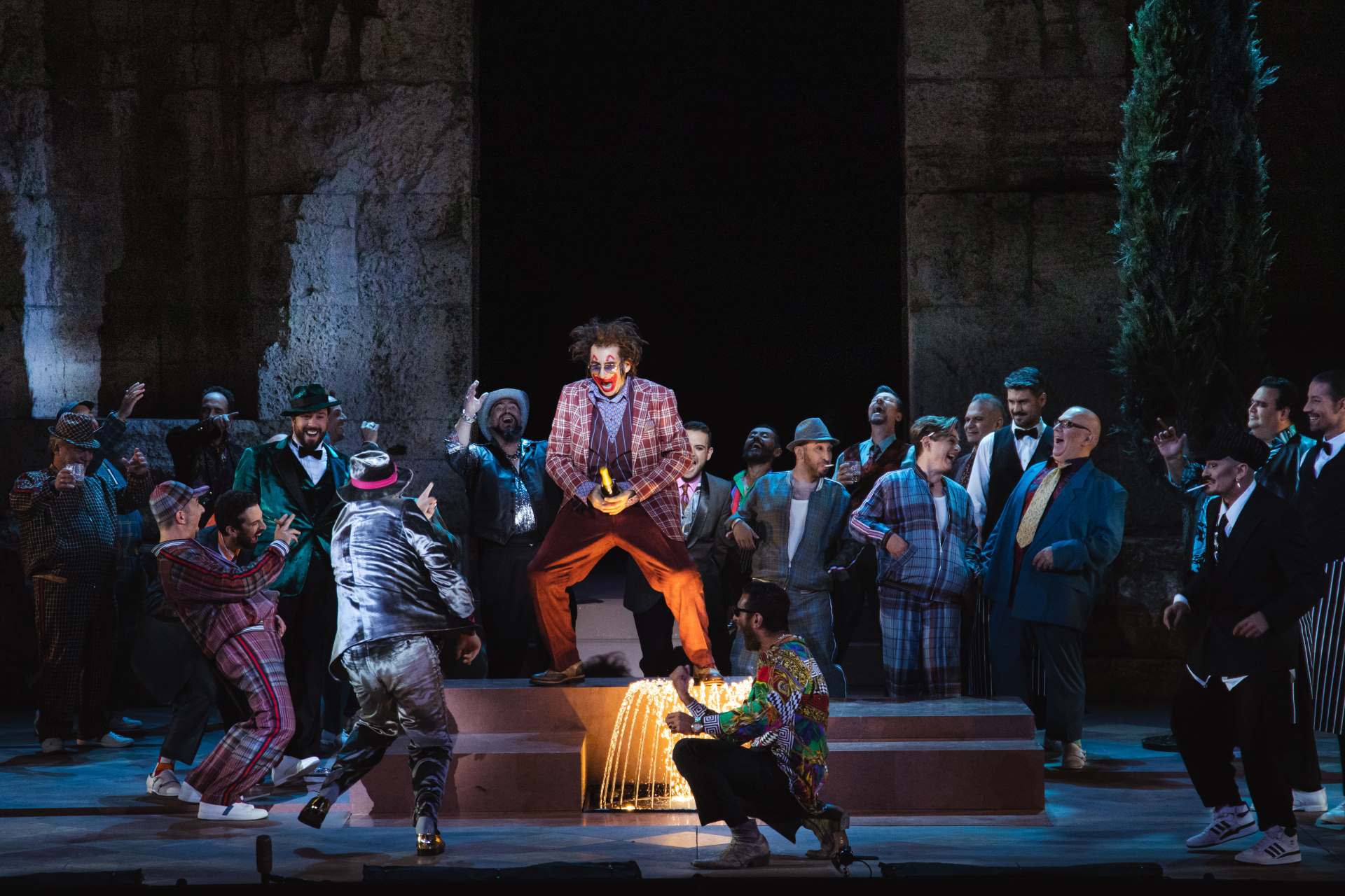 A joker dances in the middle of the stages while many other people wearing hats and colorful suits are laughing around him.