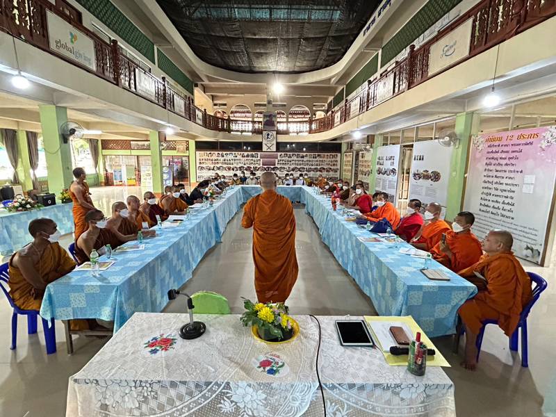 Dozens of people sit around a long U-shaped table in a large room, listening to a speaker standing at its head.