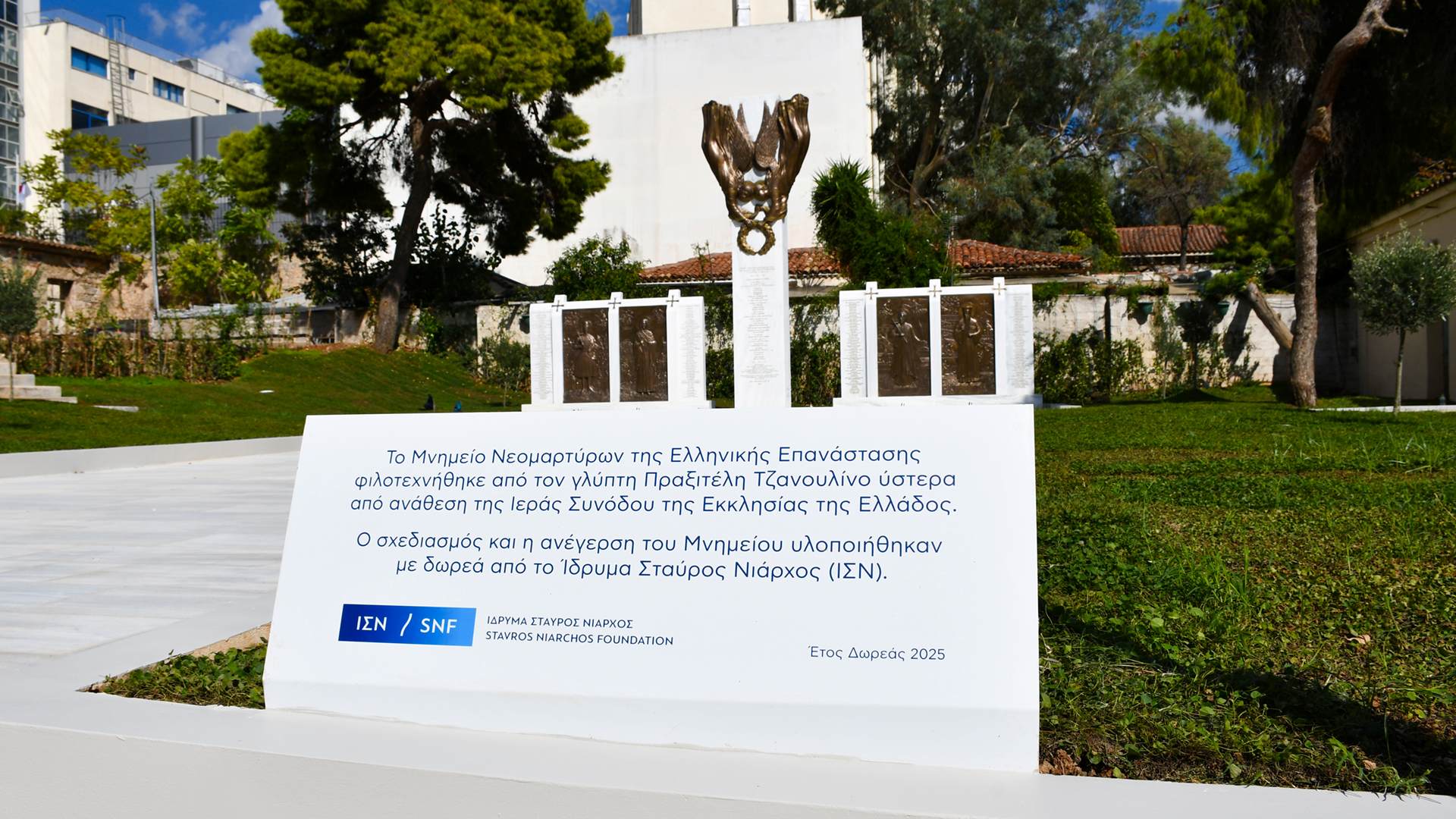 A white monument located in a green park area with a bronze reliefs on the top of it ,with engraved names on it and a big white plaque in front of it.