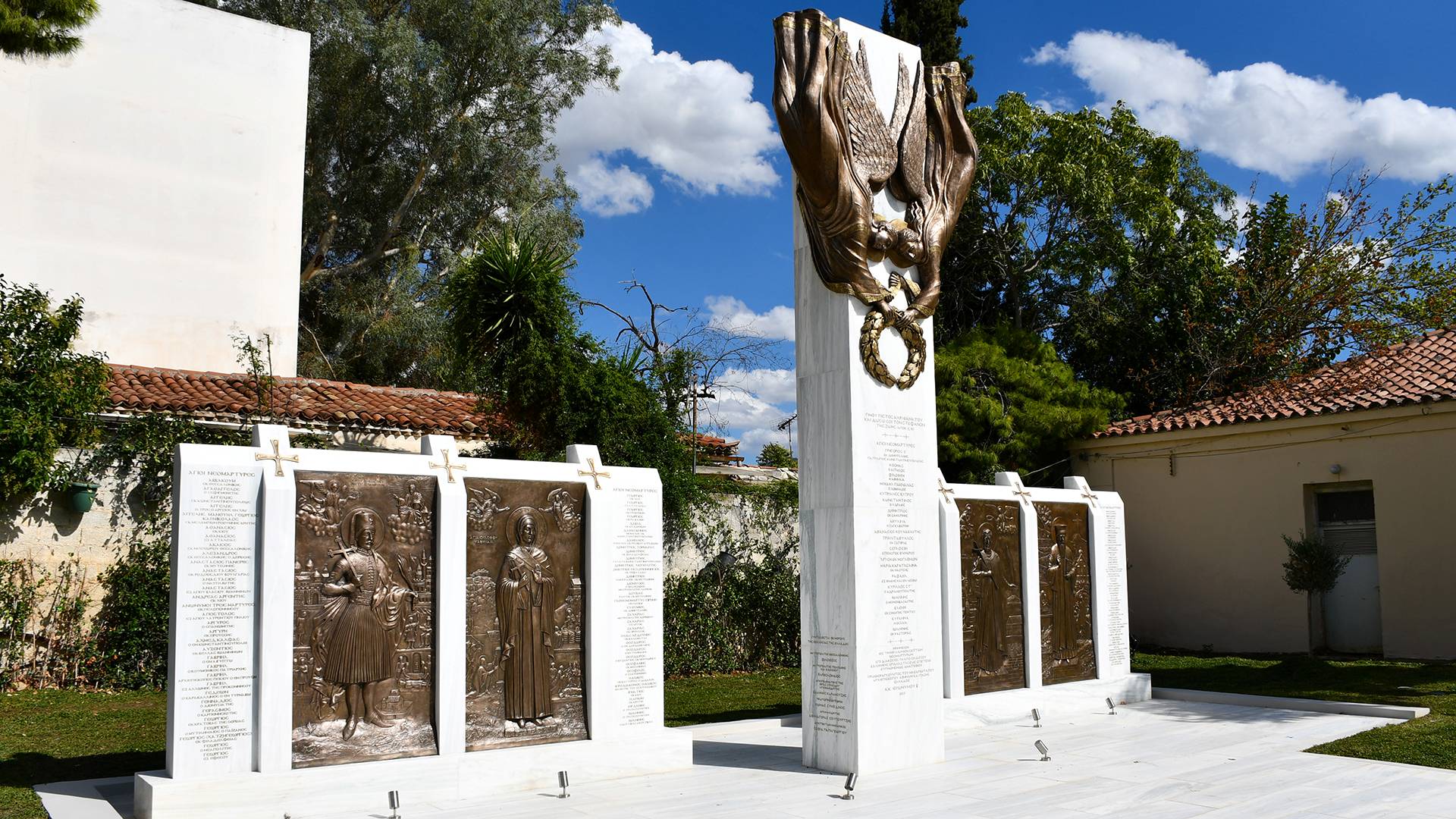 A white monument located in a green park area with a bronze reliefs on the top of it and with engraved names on it.