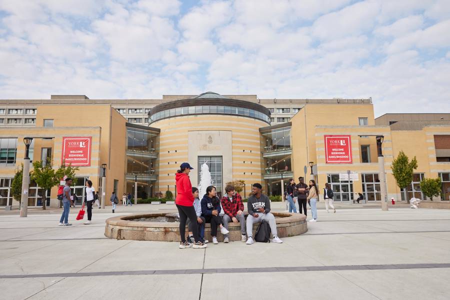 A group of students sitting on a bench outside the building of the York University, engaged in conversation and enjoying each other's company.
