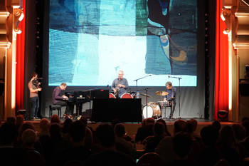 Μια μπάντα αποτελούμενη από τρεις άνδρες στο πιάνο, ντραμς και σαξόφωνο παίζουν στη σκηνή